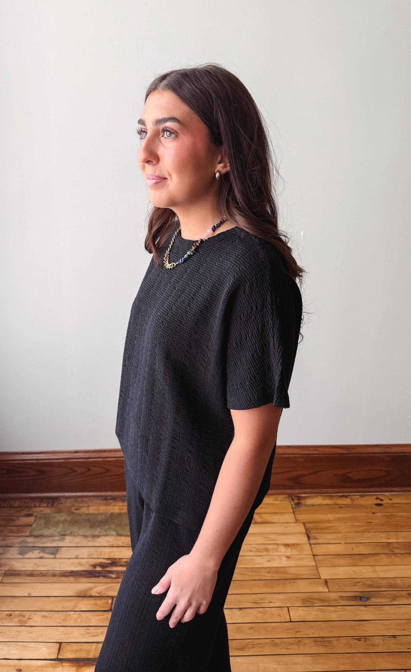 Woman wearing a black dress standing in a room with wooden flooring and a white wall.