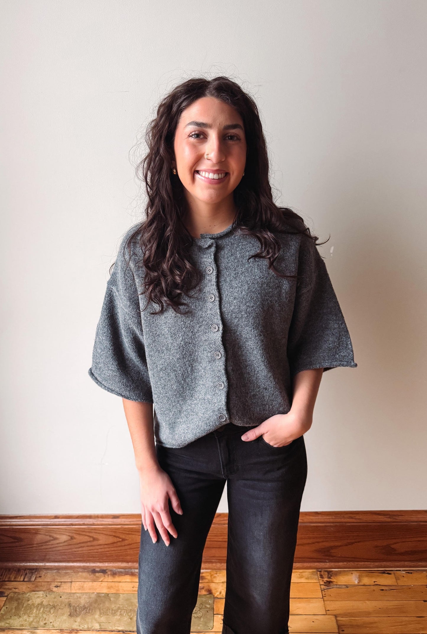 A woman with long dark curly hair stands indoors wearing a gray short-sleeve button-front cardigan or shrug and dark jeans, with one hand in her pocket. The background is a plain off-white wall with a wooden baseboard and wood floor.