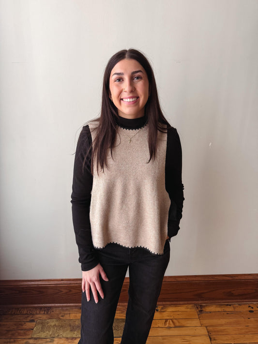 A woman with long dark hair stands indoors wearing a beige ribbed knit sweater vest over a black long-sleeve turtleneck and black pants, against a plain off-white wall with a wooden floor.