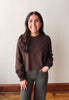 A woman with long dark hair smiling, wearing a brown knit crewneck sweater and dark olive green pants, standing against a plain off-white wall with a wooden baseboard and floor.