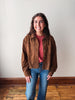 A smiling young woman stands indoors wearing a brown suede-like button-up jacket over a dusty rose top and blue jeans, posed against a plain off-white wall with a wooden floor visible.
