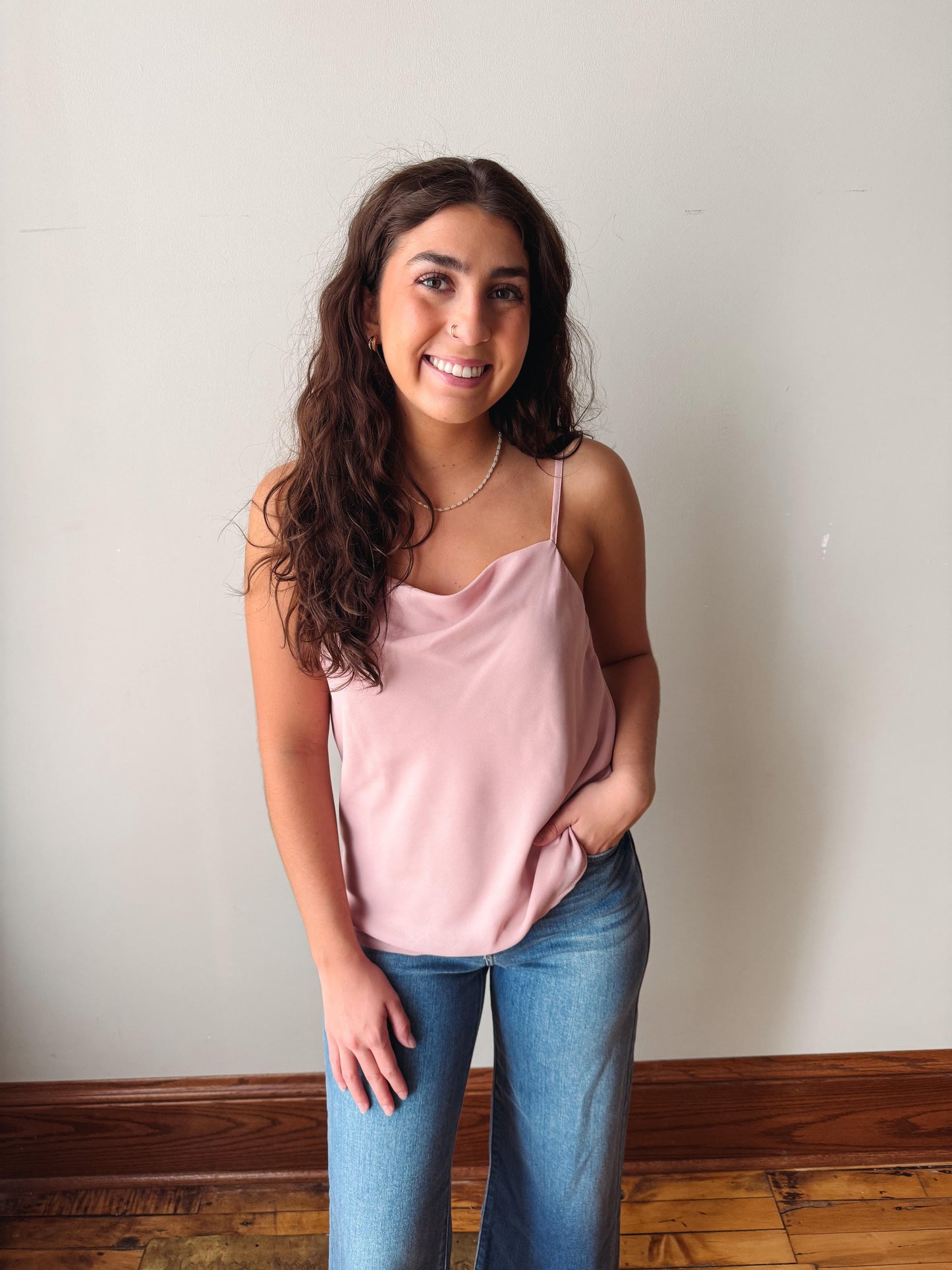 Woman wearing a pink tank top and blue jeans standing against a plain wall.
