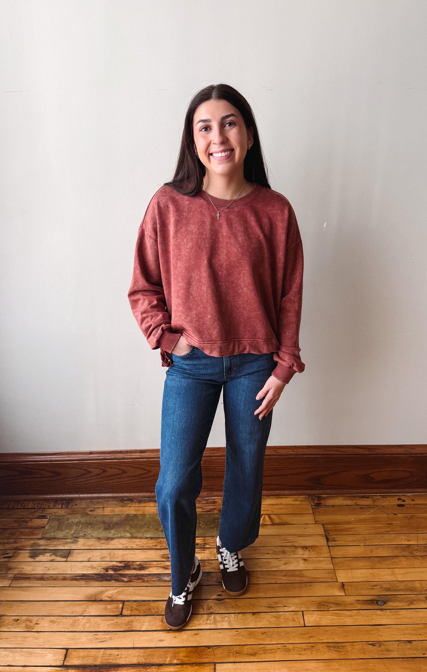A woman stands in a room with a light beige wall and wooden floor, wearing a heathered rust-red long-sleeve crewneck sweatshirt, blue jeans, and dark sneakers.