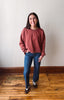 A woman stands in a room with a light beige wall and wooden floor, wearing a heathered rust-red long-sleeve crewneck sweatshirt, blue jeans, and dark sneakers.