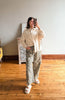 A woman takes a mirror selfie in a room with hardwood floors, wearing a cream-colored long-sleeve button-down shirt and wide-leg patterned pants with white slide sandals.