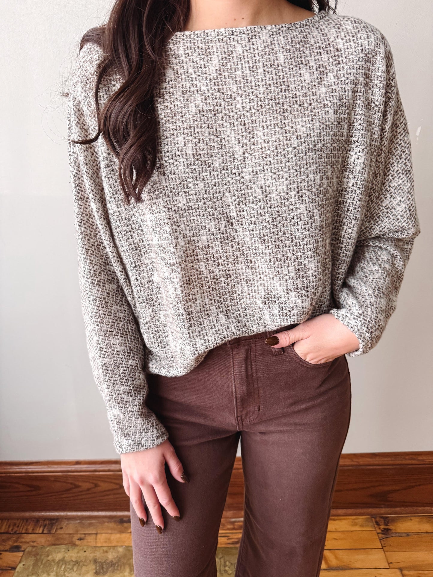 A woman wearing a textured, long-sleeve knit sweater in a heathered gray and white pattern, paired with brown pants, standing indoors against a plain wall.