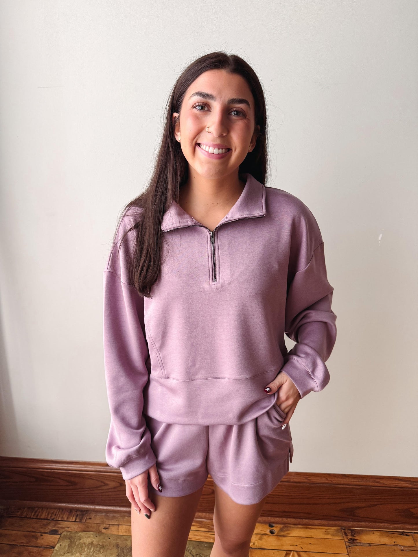A woman modeling a matching dusty purple lounge set consisting of a quarter-zip sweatshirt and matching shorts, standing against a plain off-white wall with wooden flooring visible at the bottom.