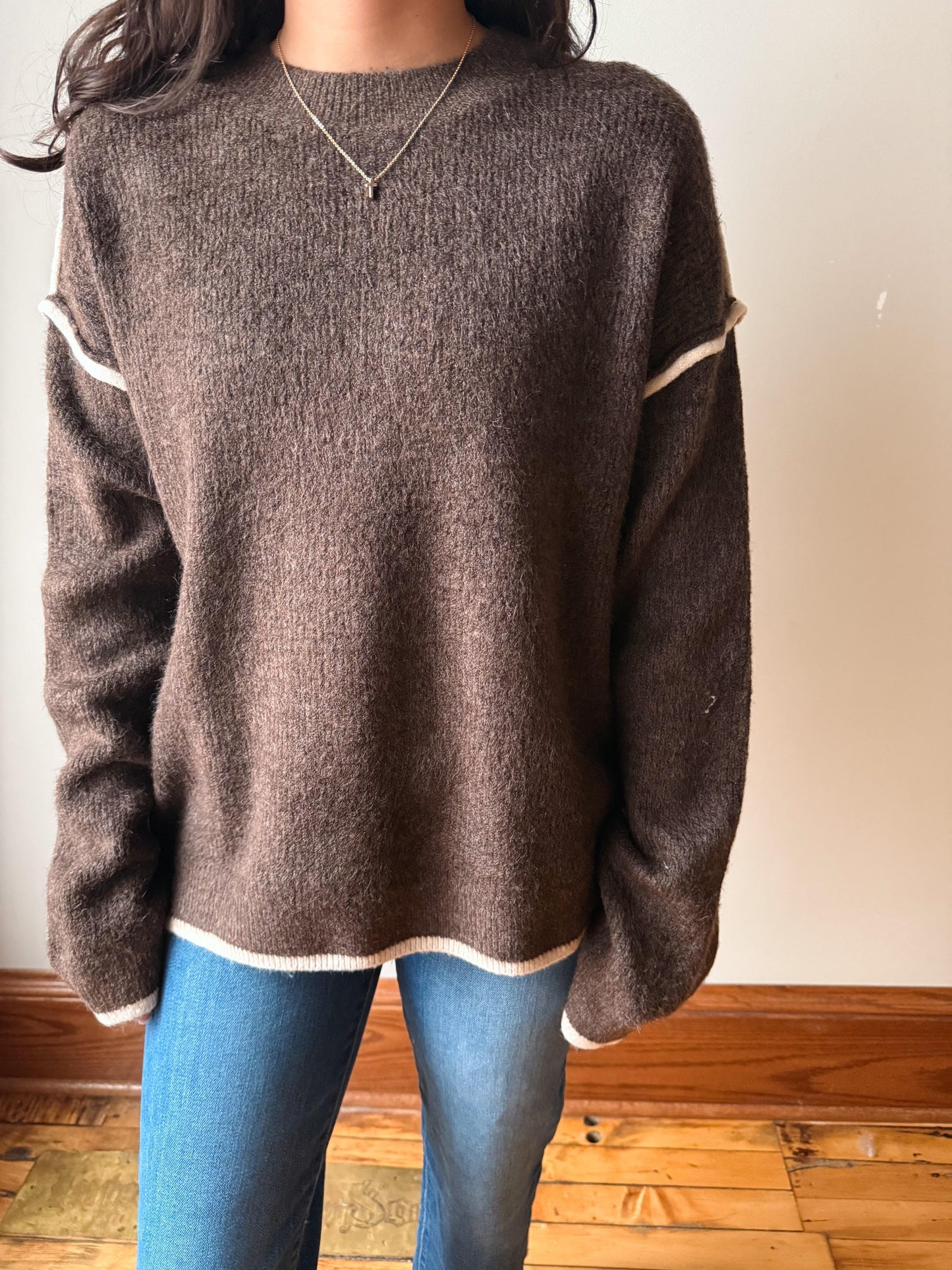 A person wearing a brown fuzzy knit sweater with cream-colored trim along the collar, cuffs, and hem, paired with blue jeans, standing indoors against a plain wall and wooden floor.