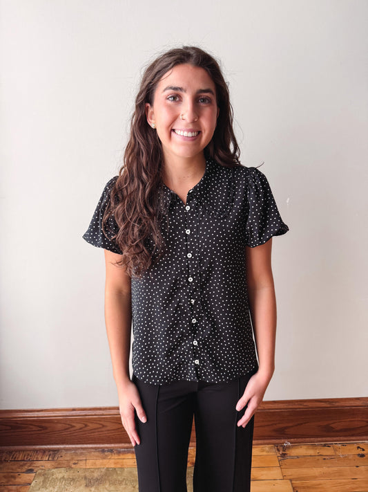 A smiling young woman with long brown hair stands indoors wearing a black short-sleeve button-up shirt with a white polka dot pattern and black pants.