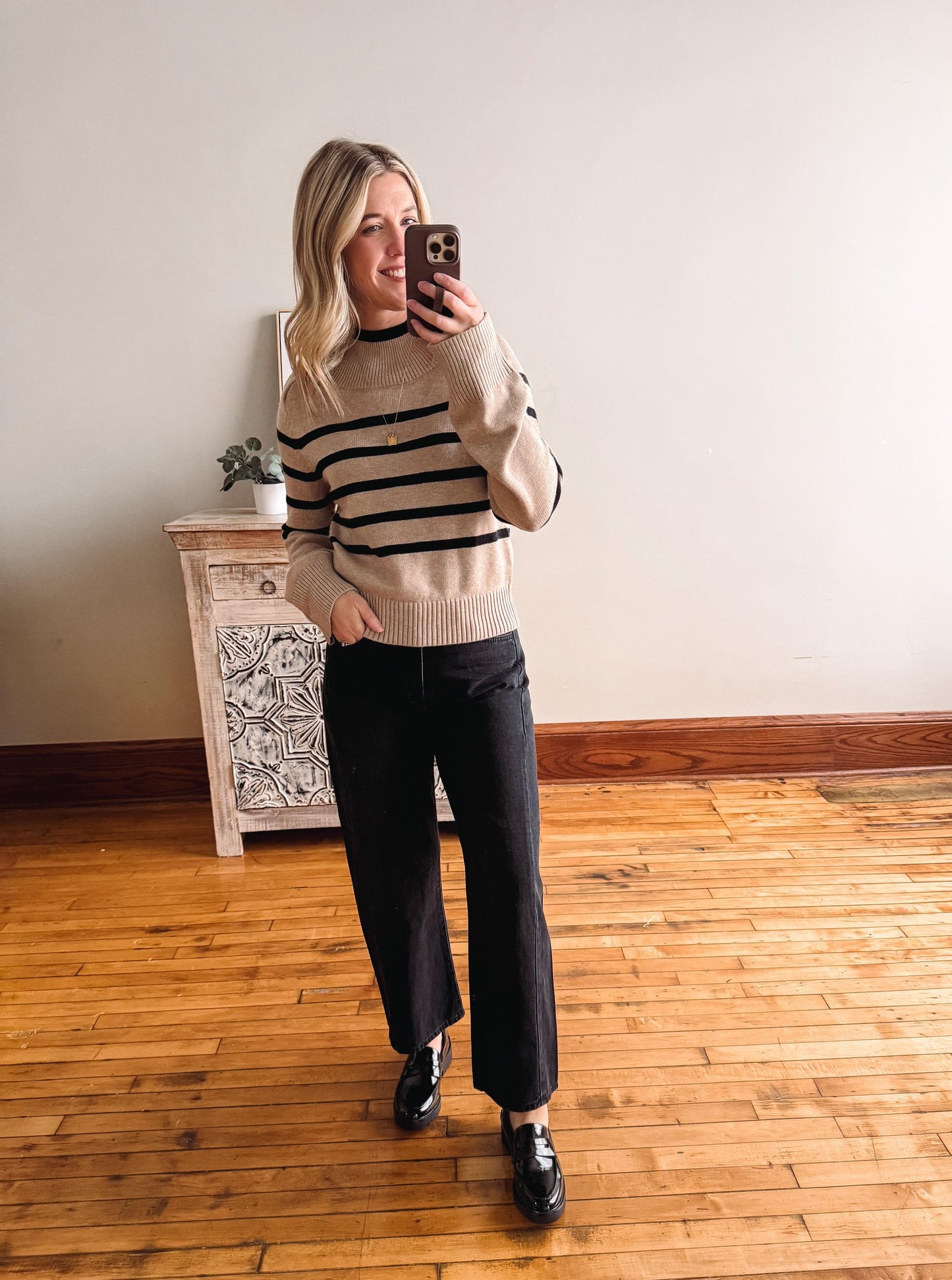 Person wearing a beige sweater with black stripes and dark pants, standing in a room with wooden flooring and a decorative cabinet.