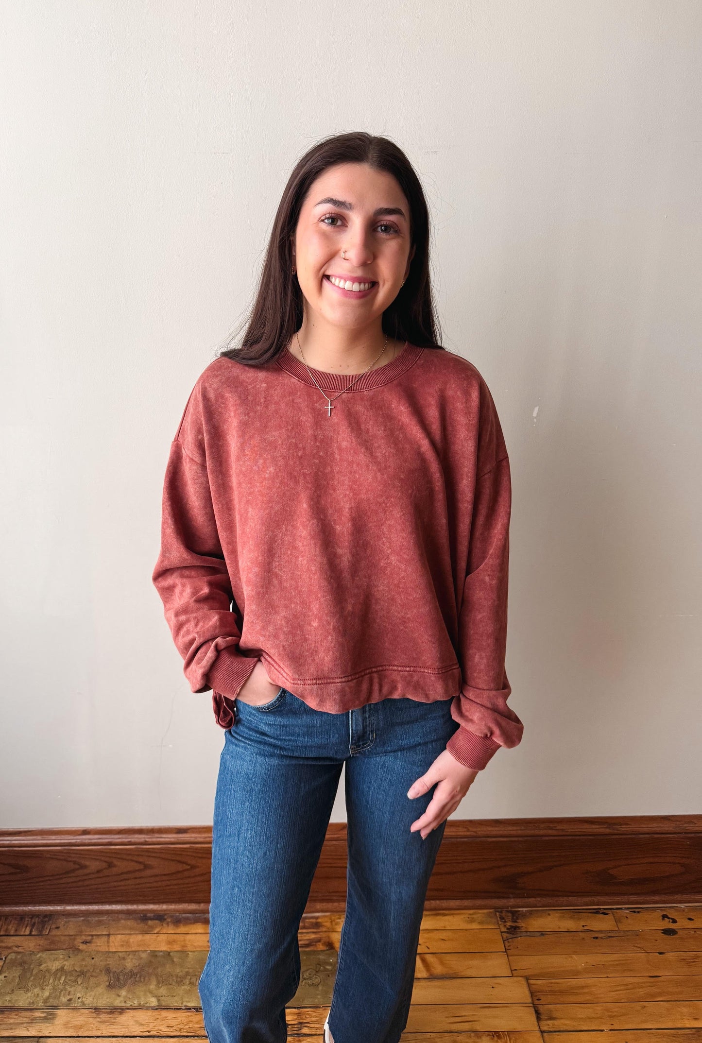 A woman wearing a washed, heathered dusty rose crewneck sweatshirt and blue jeans, standing against a plain off-white wall with a wooden floor visible.
