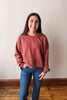 A woman wearing a washed, heathered dusty rose crewneck sweatshirt and blue jeans, standing against a plain off-white wall with a wooden floor visible.