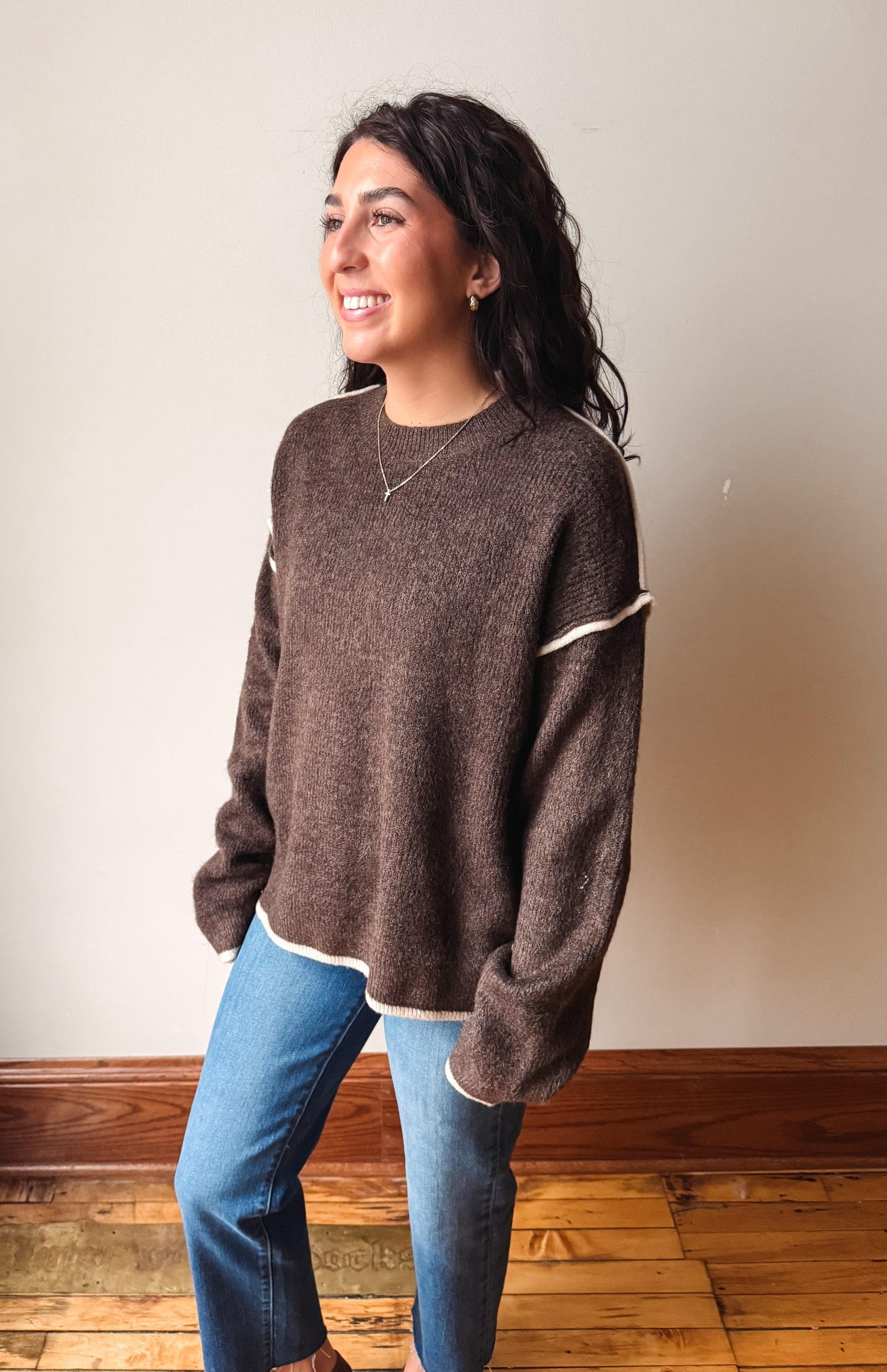 A woman wearing a brown knit sweater with cream-colored contrast trim on the shoulders and hem, paired with blue jeans, standing in a room with a beige wall and wooden floor.