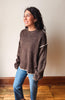 A woman wearing a brown knit sweater with cream-colored contrast trim on the shoulders and hem, paired with blue jeans, standing in a room with a beige wall and wooden floor.