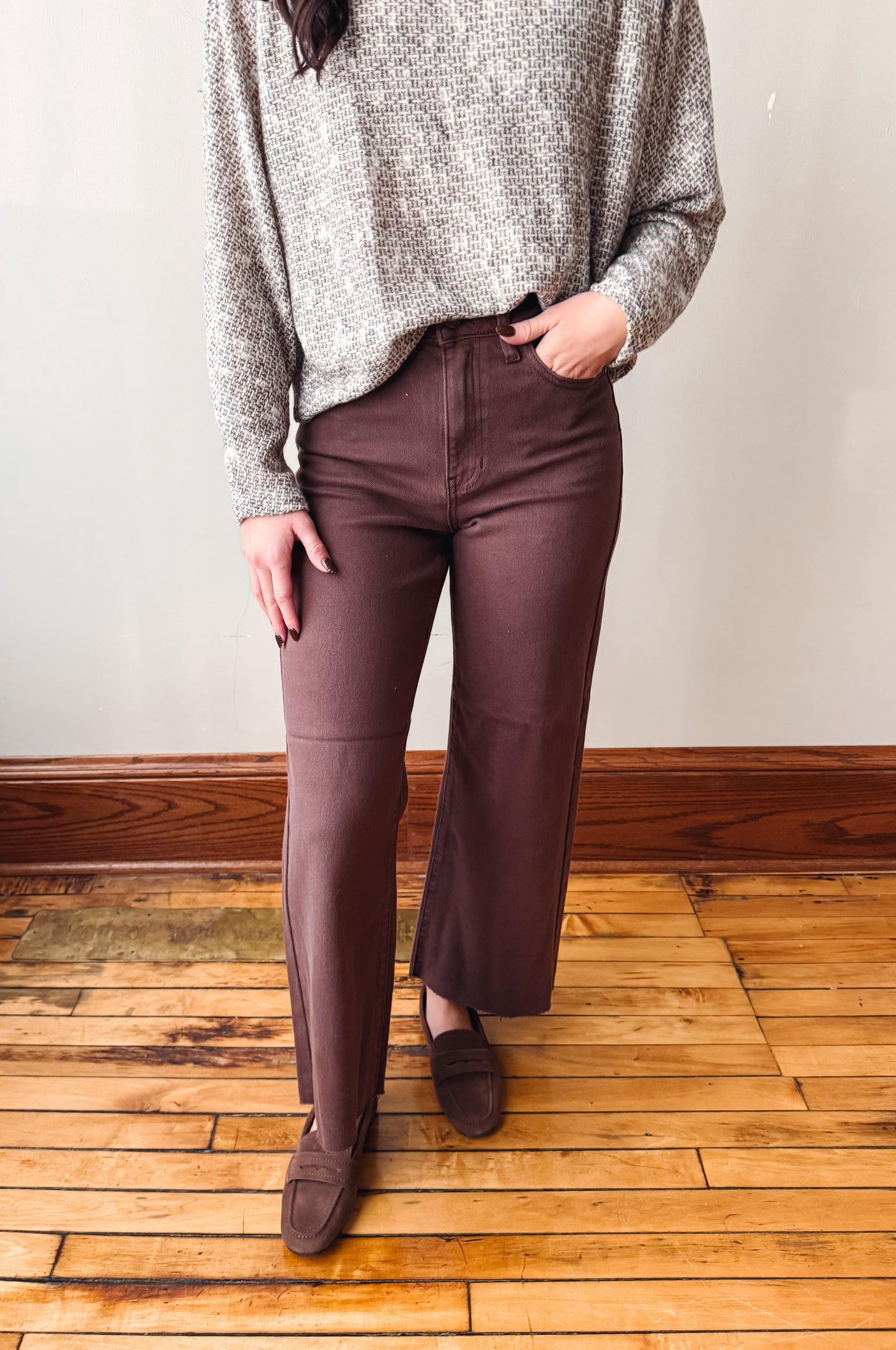 A person wearing brown wide-leg cropped pants, a gray knit sweater, and brown loafers, standing on a wooden floor against a plain wall.