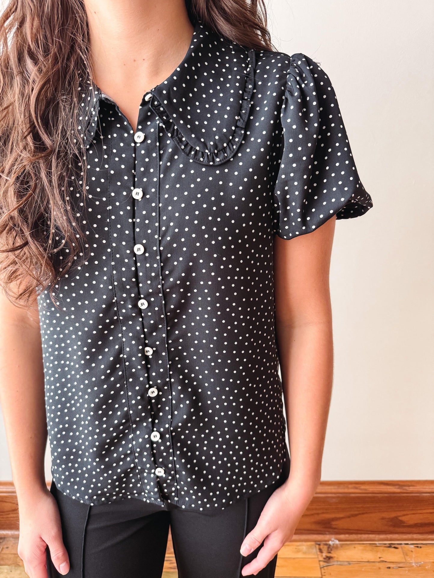 A woman wearing a black short-sleeve button-up blouse with a white polka dot pattern and ruffled collar, paired with black pants.