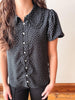 A woman wearing a black short-sleeve button-up blouse with a white polka dot pattern and ruffled collar, paired with black pants.
