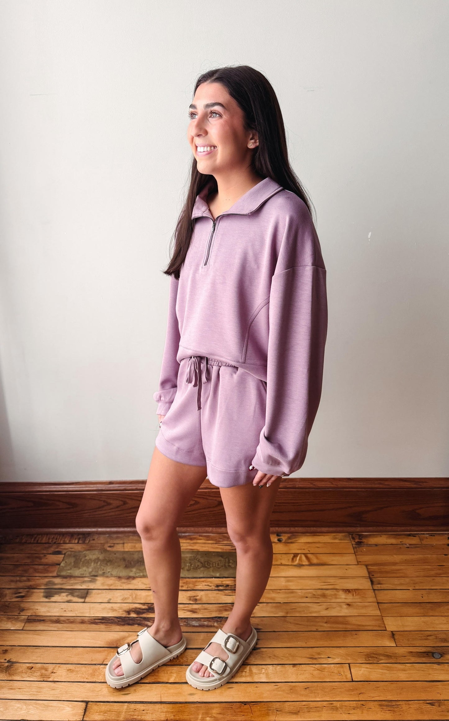 A woman modeling a matching lavender lounge set consisting of a half-zip long-sleeve sweatshirt and matching shorts, paired with beige slide sandals, standing on a wooden floor against a plain wall.