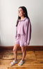 A woman modeling a matching lavender lounge set consisting of a half-zip long-sleeve sweatshirt and matching shorts, paired with beige slide sandals, standing on a wooden floor against a plain wall.