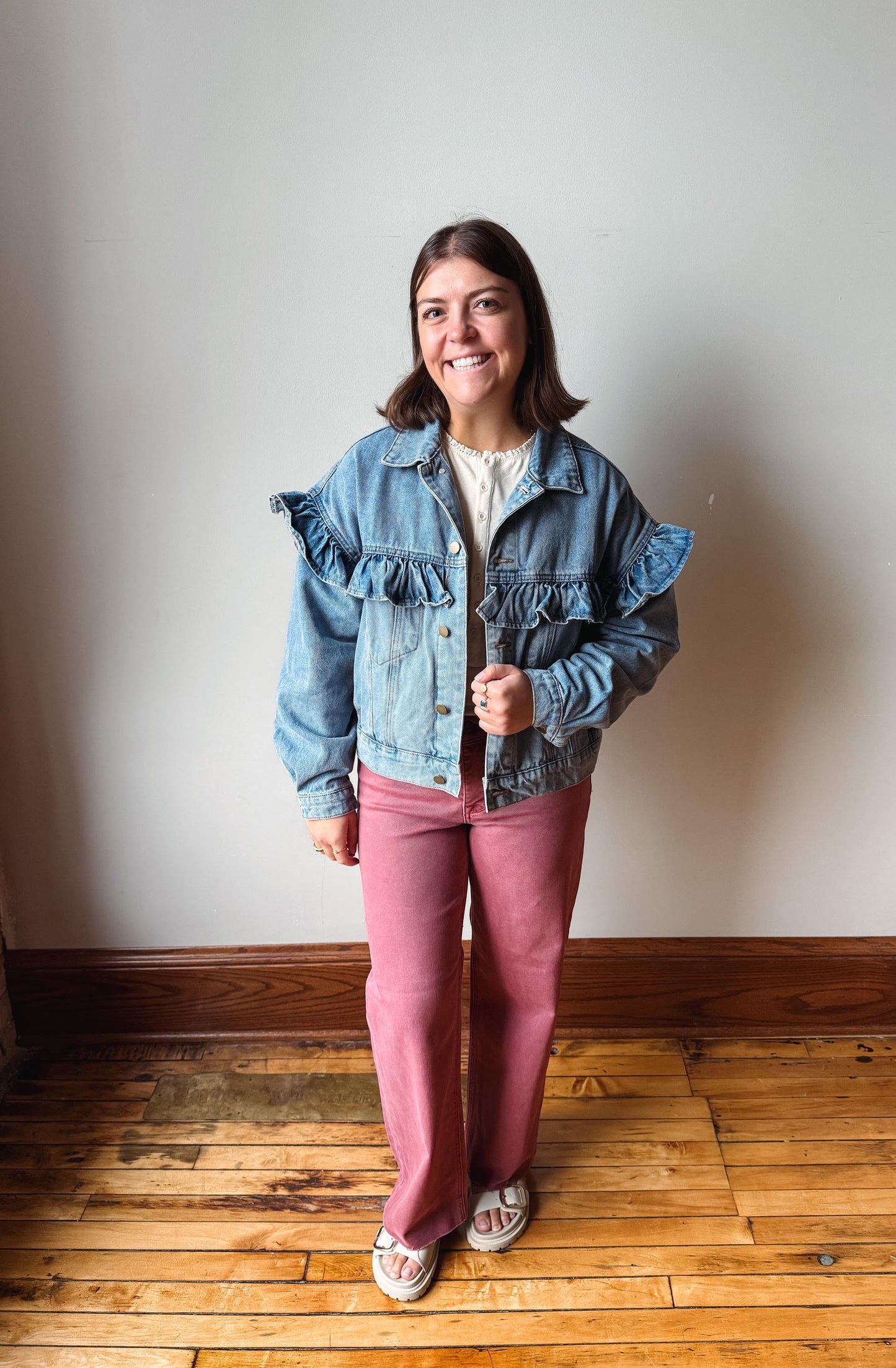 A woman stands on a wooden floor against a plain wall, wearing a light blue denim jacket with ruffle details on the shoulders and sleeves, paired with dusty rose pants and white sneakers.
