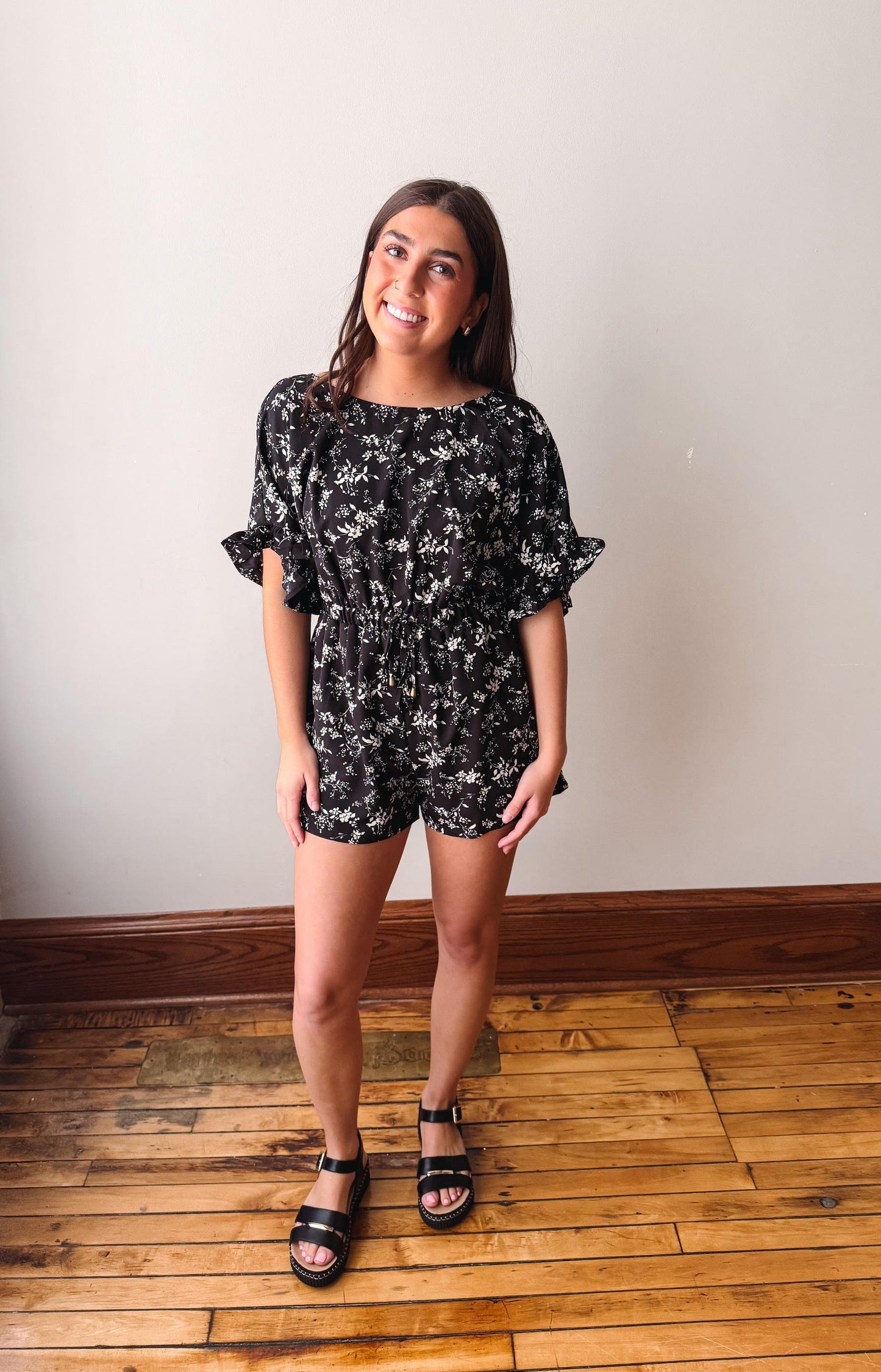 Woman wearing a black floral romper standing on a wooden floor with a white wall background