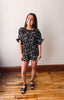 Woman wearing a black floral romper standing on a wooden floor with a white wall background