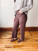 A person wearing wide-leg mauve-brown corduroy pants, a gray knit sweater, and brown suede loafers, standing on a wooden floor against a white wall.