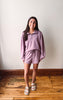 A woman modeling a matching lavender lounge set consisting of a long-sleeve quarter-zip sweatshirt and coordinating shorts, paired with white slide sandals, standing on a wooden floor against a plain off-white wall.