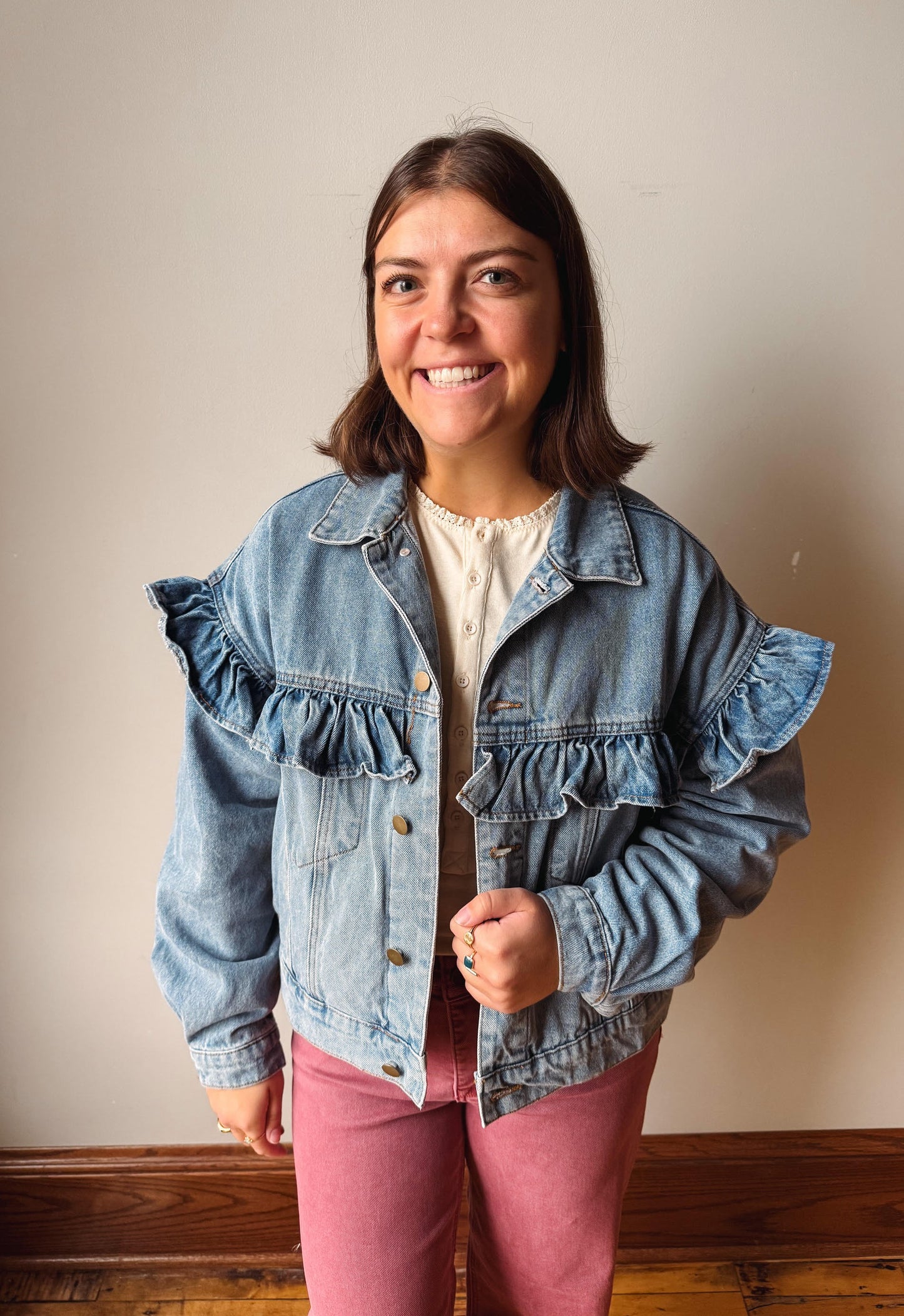 A smiling woman wearing a light blue denim jacket with dramatic ruffle details on the shoulders and front, paired with a cream top and dusty pink pants, standing against a plain beige wall.