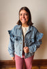 A smiling woman wearing a light blue denim jacket with dramatic ruffle details on the shoulders and front, paired with a cream top and dusty pink pants, standing against a plain beige wall.