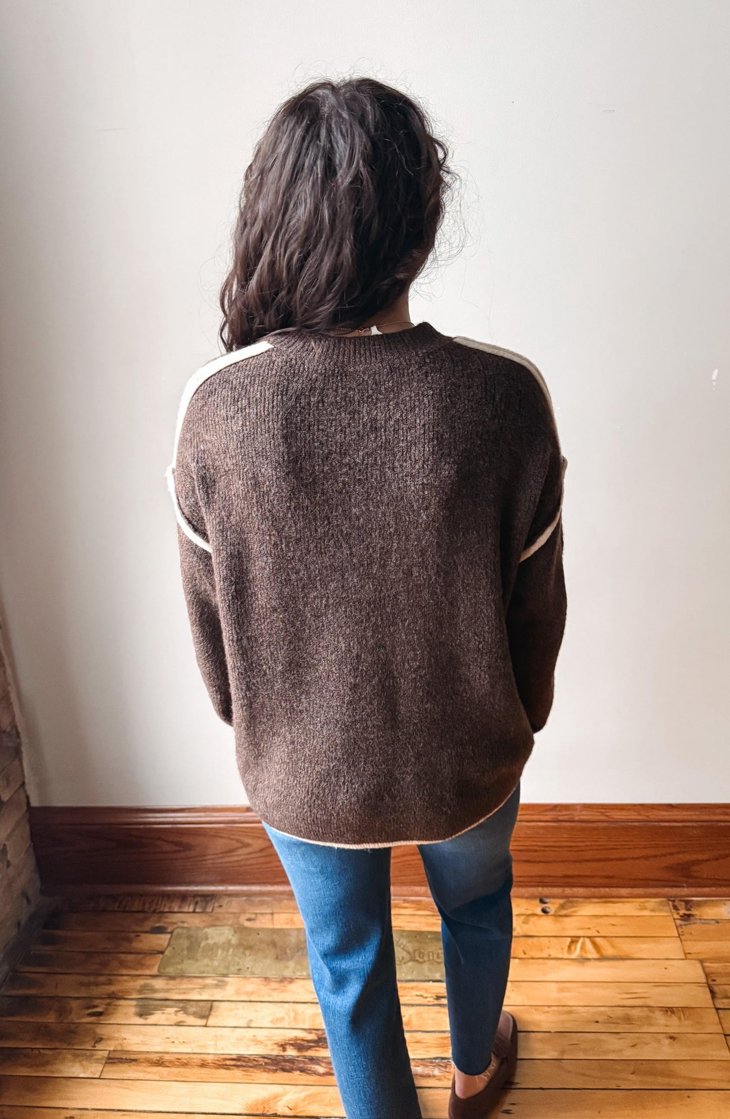 A person viewed from behind wearing a brown knit sweater with cream-colored shoulder stripes, paired with blue jeans, standing on a wooden floor against a plain off-white wall.