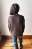 A person viewed from behind wearing a brown knit sweater with cream-colored shoulder stripes, paired with blue jeans, standing on a wooden floor against a plain off-white wall.