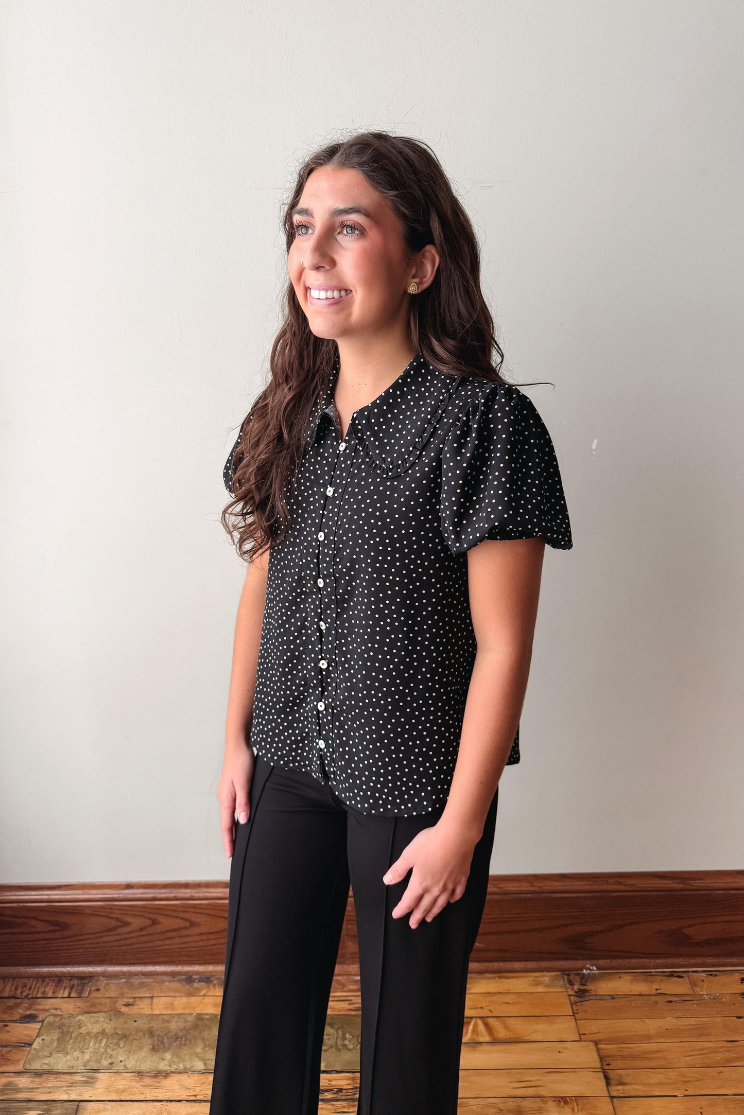 A woman modeling a black short-sleeve button-up shirt with a white polka dot pattern, paired with black pants, standing in front of a plain off-white wall with a wooden floor visible.