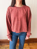 A person wearing a washed, faded dusty rose crewneck sweatshirt and blue jeans, standing against a plain off-white wall with a wooden baseboard and floor.