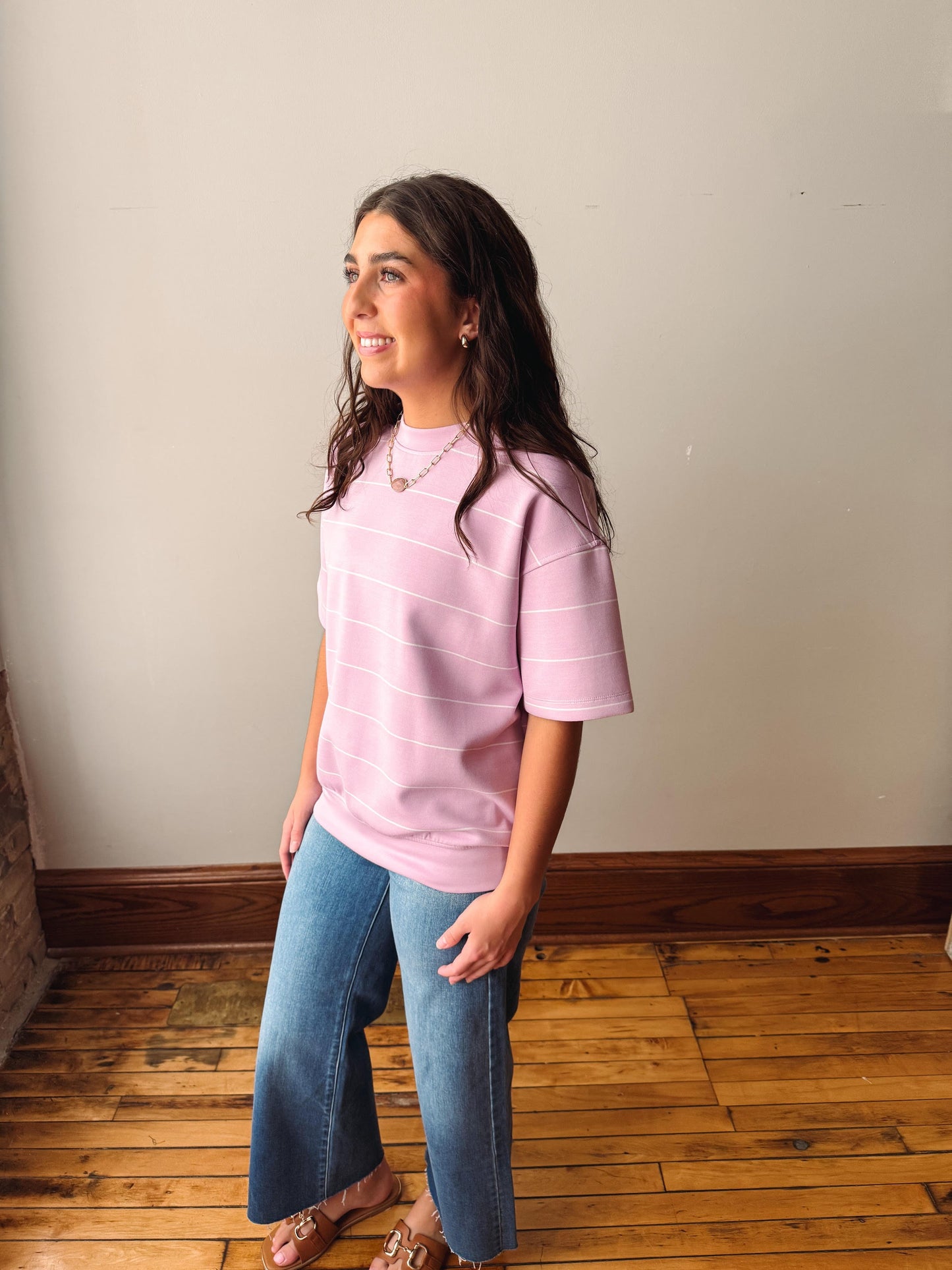 Woman wearing a pink shirt and blue jeans standing in a room with wooden flooring and a white wall.