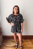 Woman wearing a black floral romper standing in a room with wooden flooring and a white wall.