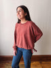 A woman modeling a washed, dusty rose long-sleeve crewneck sweatshirt with side tie details, paired with blue jeans, standing against a plain off-white wall.