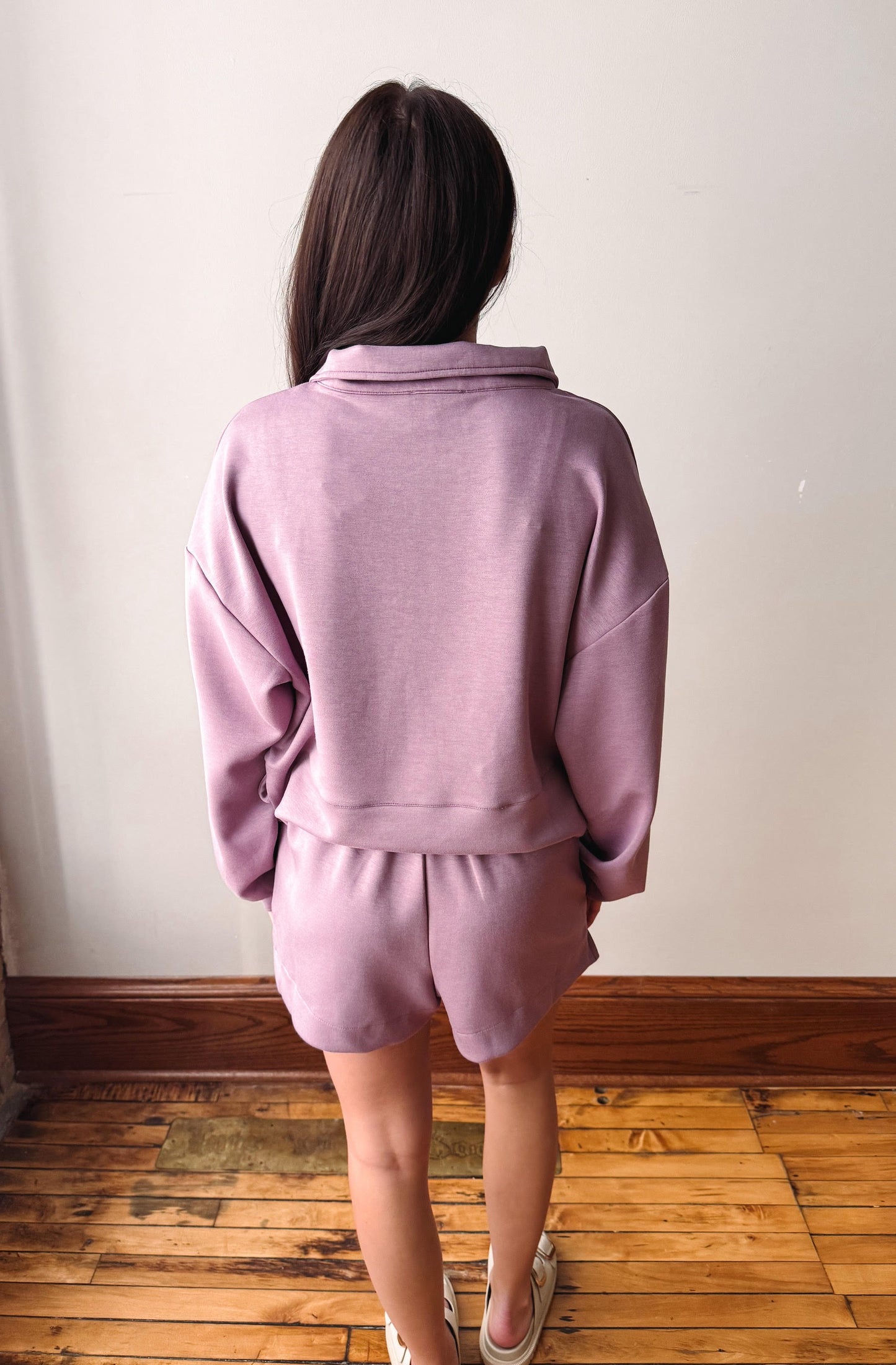A person viewed from behind wearing a matching lavender sweatshirt and shorts set, standing on a wooden floor against a plain off-white wall.