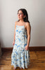 Woman wearing a floral dress standing on a wooden floor against a plain wall.