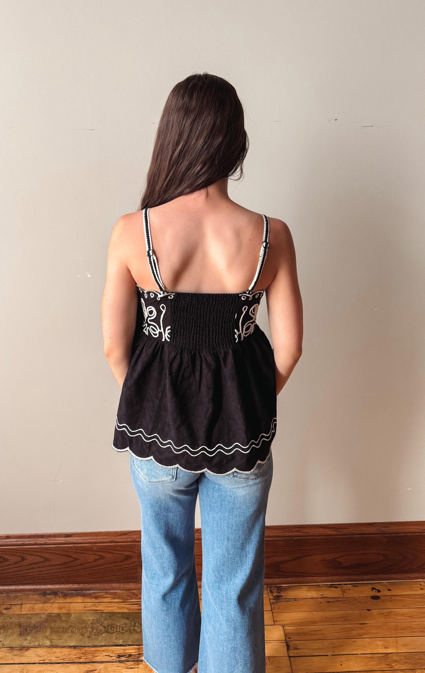 Person wearing a black top with white embroidery and blue jeans against a plain wall.