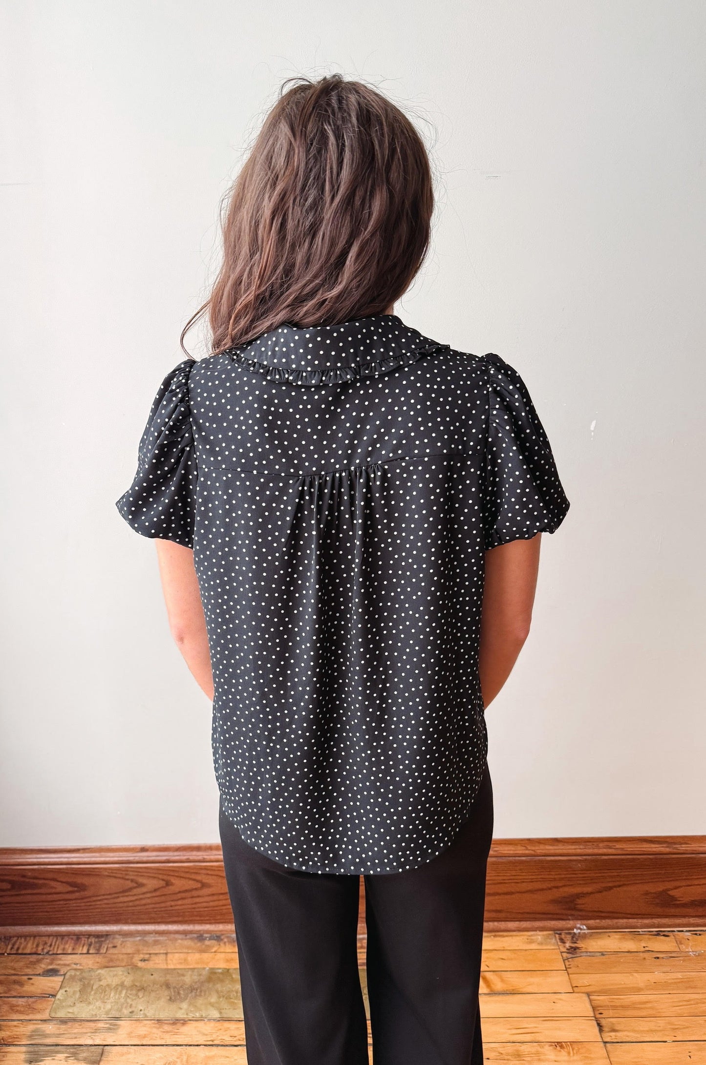 A person viewed from behind wearing a black short-sleeve blouse with white polka dots and black pants, standing in front of a plain off-white wall with a wooden baseboard and wood floor.