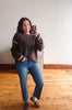 A woman takes a mirror selfie wearing a dark brown knit sweater with white stripe accents on the sleeves, blue jeans, and brown loafers, standing in a room with a light wall and wooden floor.