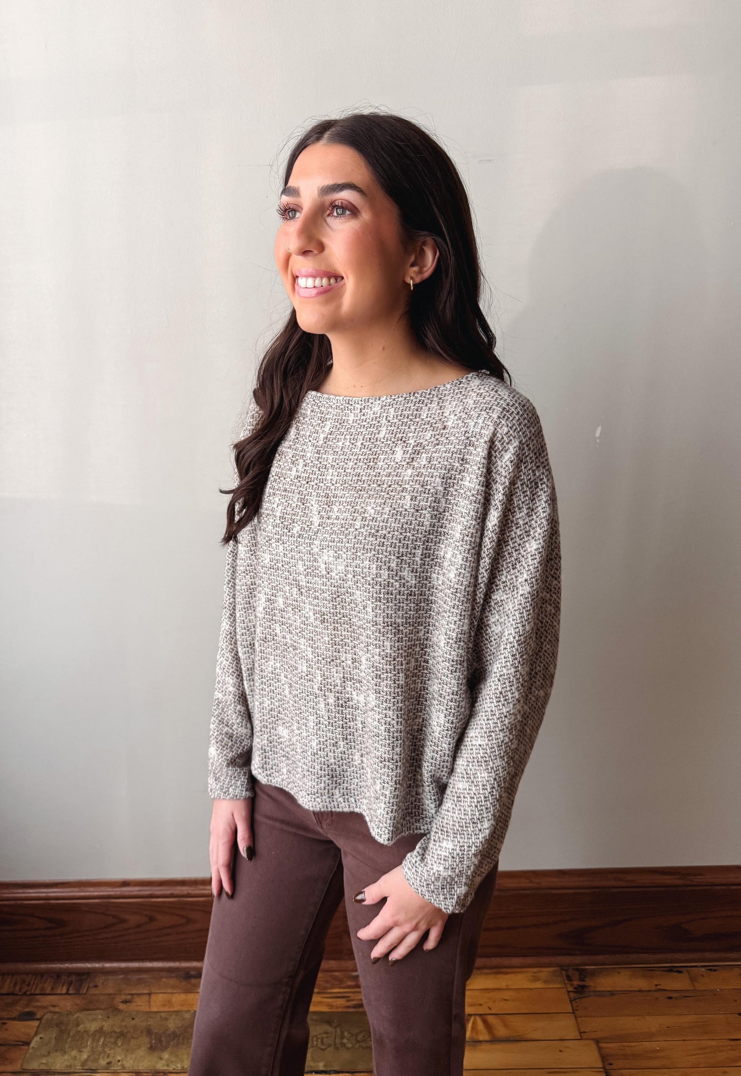 A woman wearing a textured, heathered gray and white knit long-sleeve sweater and brown pants, standing in an indoor setting with a light gray wall and wooden floor.