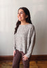 A woman wearing a textured, heathered gray and white knit long-sleeve sweater and brown pants, standing in an indoor setting with a light gray wall and wooden floor.