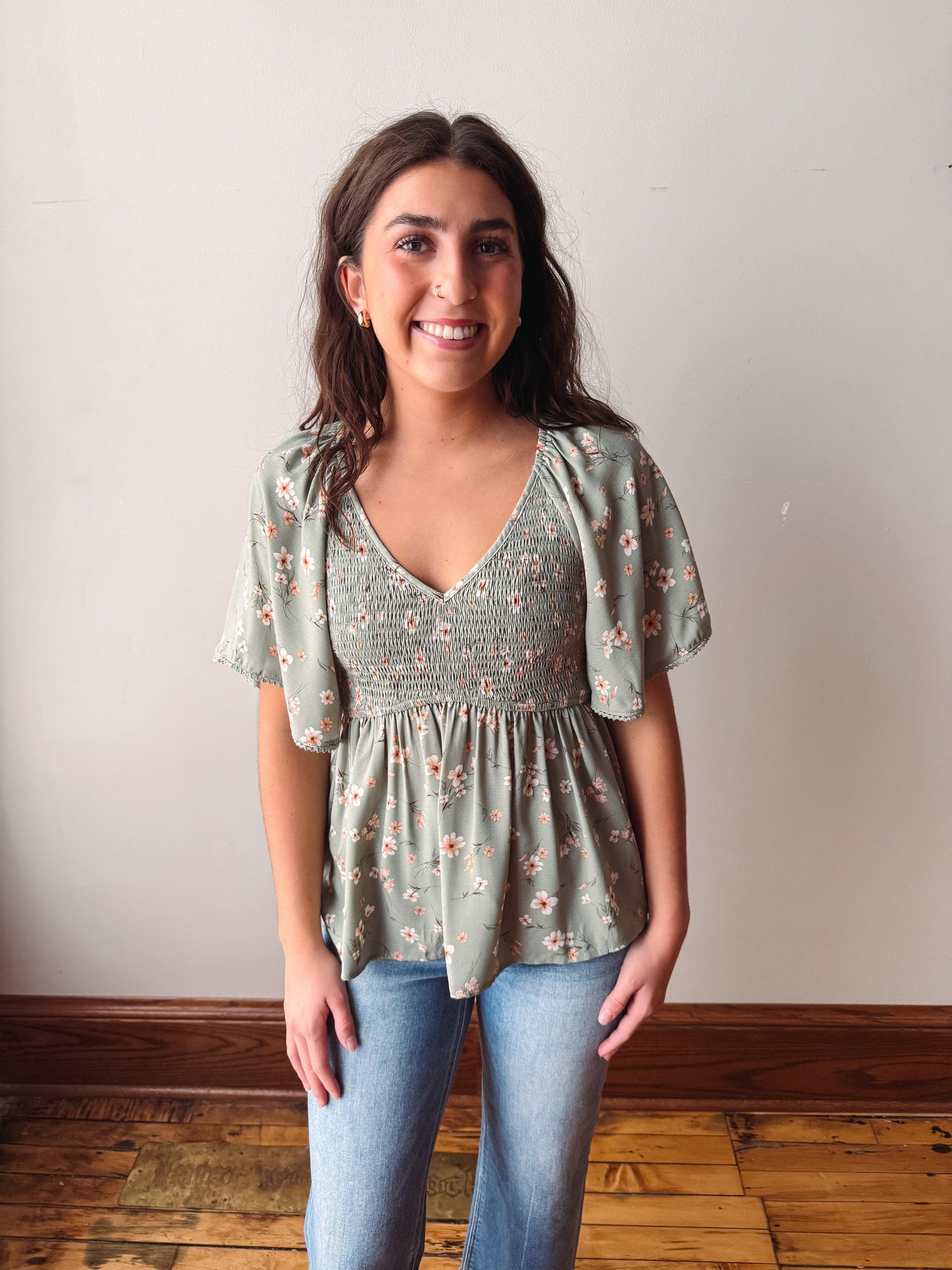 Woman wearing a floral blouse and jeans against a plain wall.