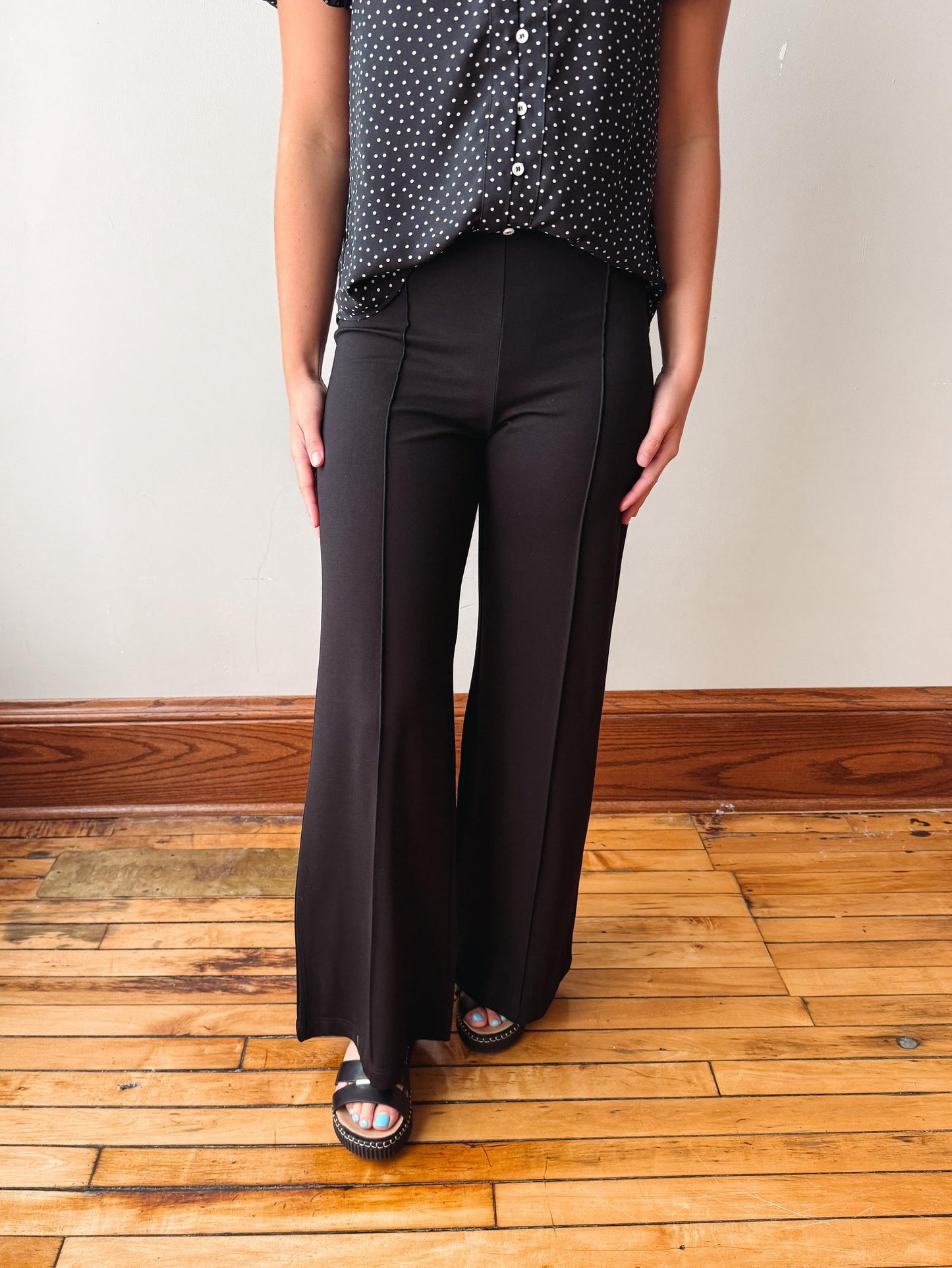 A person wearing black wide-leg pants with a black and white polka dot button-up shirt, standing on a wooden floor against a plain wall.