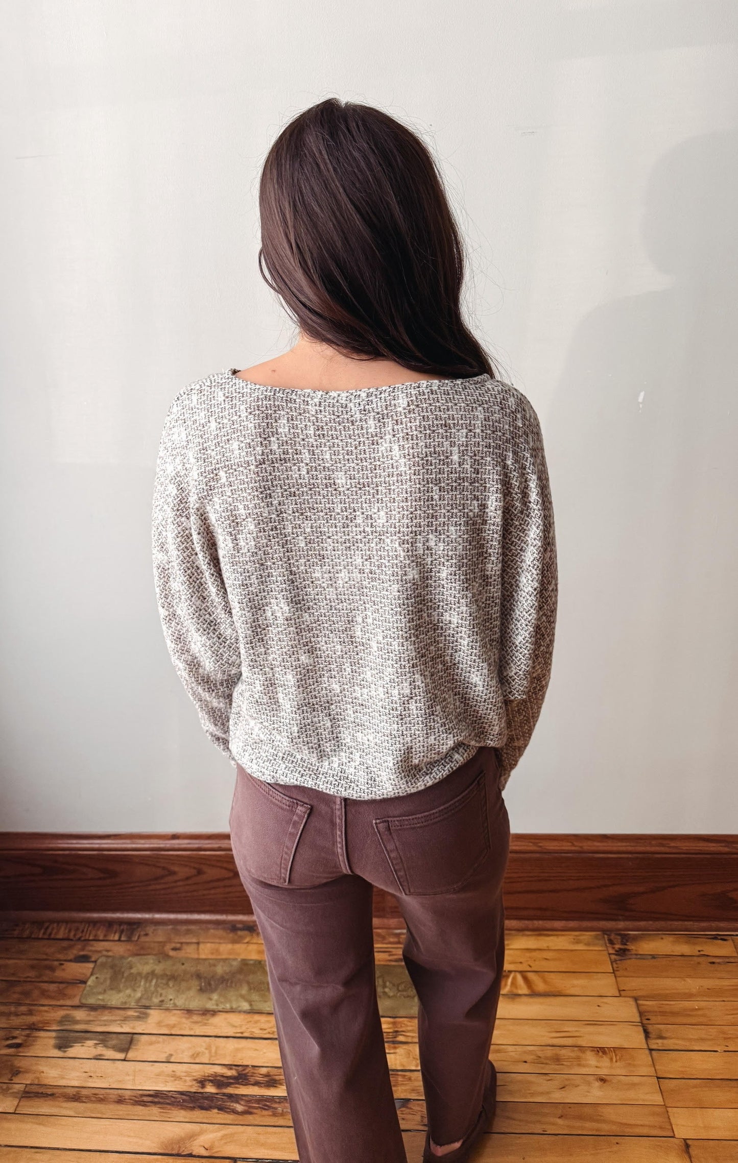 A woman viewed from behind wearing a textured, speckled light gray knit sweater and mauve-brown pants, standing on a wooden floor against a plain off-white wall.