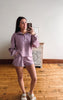 A woman takes a mirror selfie wearing a matching lavender purple lounge set consisting of a quarter-zip long-sleeve top and matching shorts, standing on a wooden floor in front of a white wall.