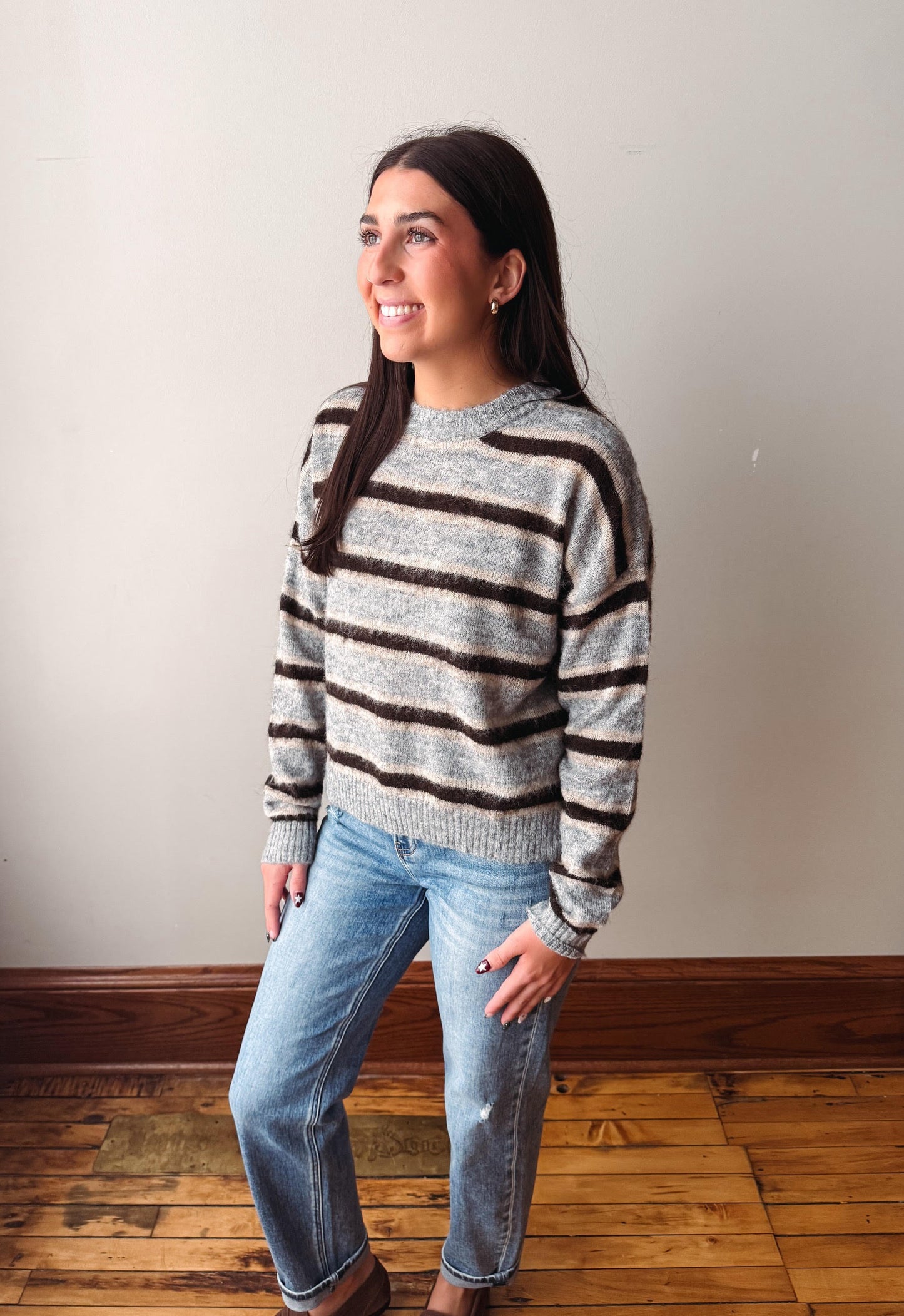 A woman wearing a gray and brown horizontally striped knit sweater and light blue jeans, standing on a wooden floor against a plain off-white wall.