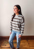 A woman wearing a gray and brown horizontally striped knit sweater and light blue jeans, standing on a wooden floor against a plain off-white wall.